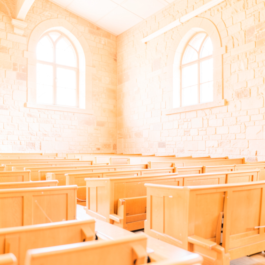 Bright, airy photograph of a grand historic auditorium interior with warm stone walls, arched windows, and rows of wooden seating bathed in natural light