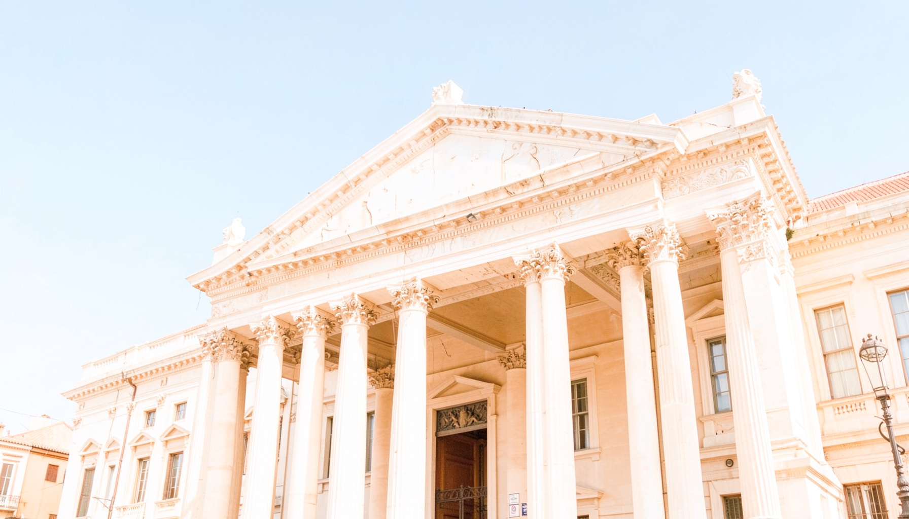 Wide, luminous photograph of a stately Roman building facade with classical columns and a clear blue sky, conveying a sense of history and elegance