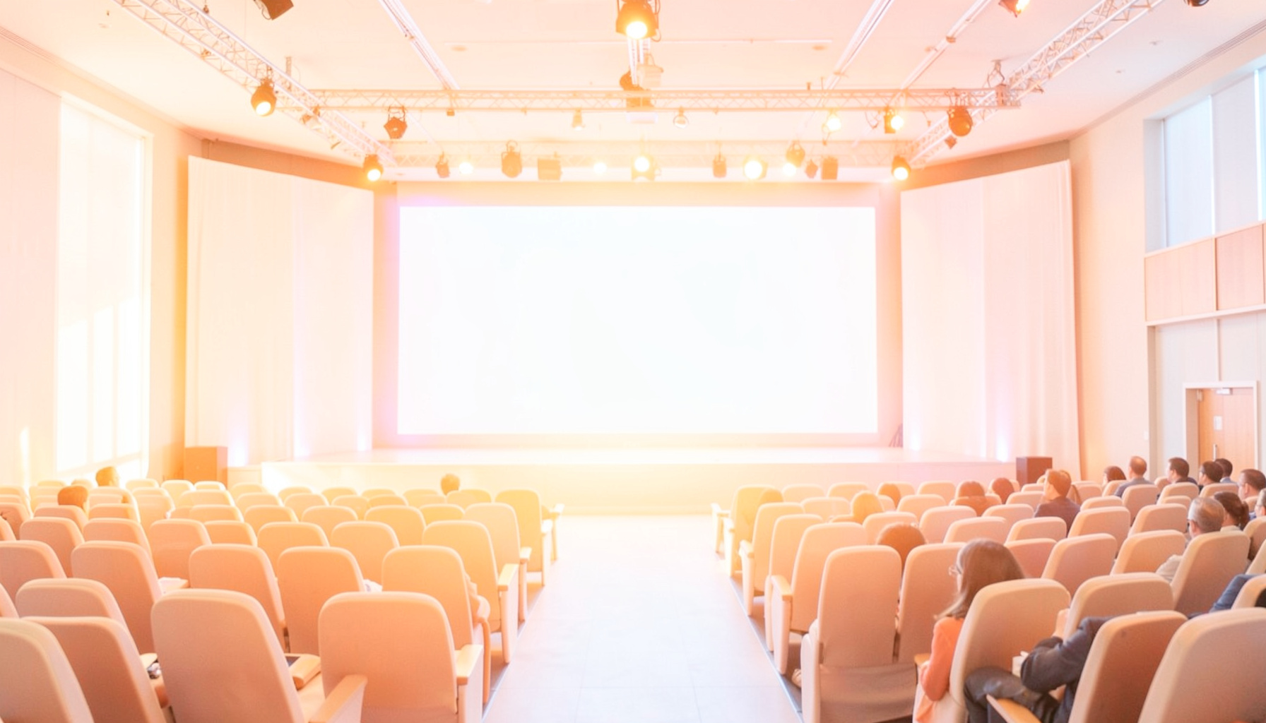 A bright conference stage with a large projection screen and rows of empty audience seating, bathed in warm amber and soft blue lighting.