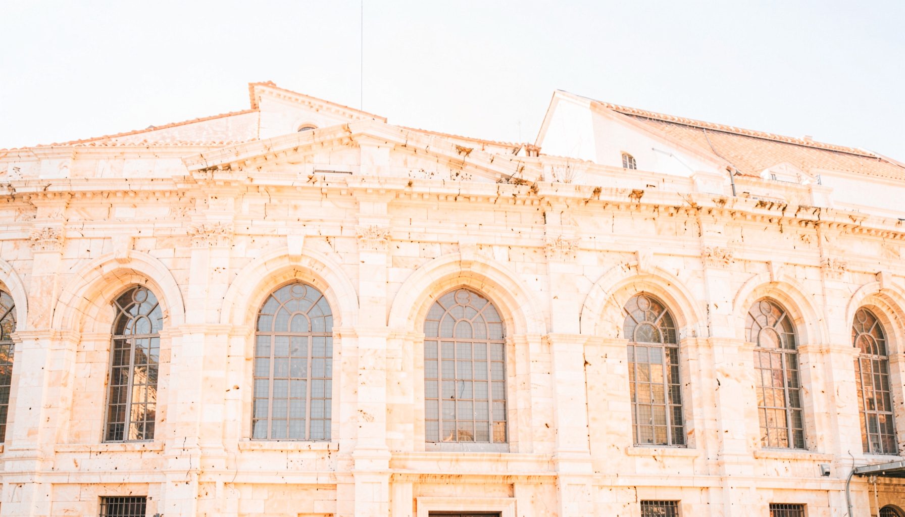 Warm stone facade of a historic Roman auditorium with tall arched windows, bathed in soft afternoon light against a clear sky.