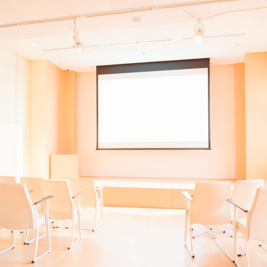 A bright, high-key photograph of a modern conference presentation setup with a large screen and a softly lit stage, warm neutral tones.